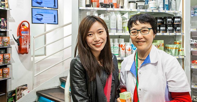 Deux personnes sourient dans une pharmacie, debout près d'étagères de médicaments et d'un extincteur rouge fixé au mur.