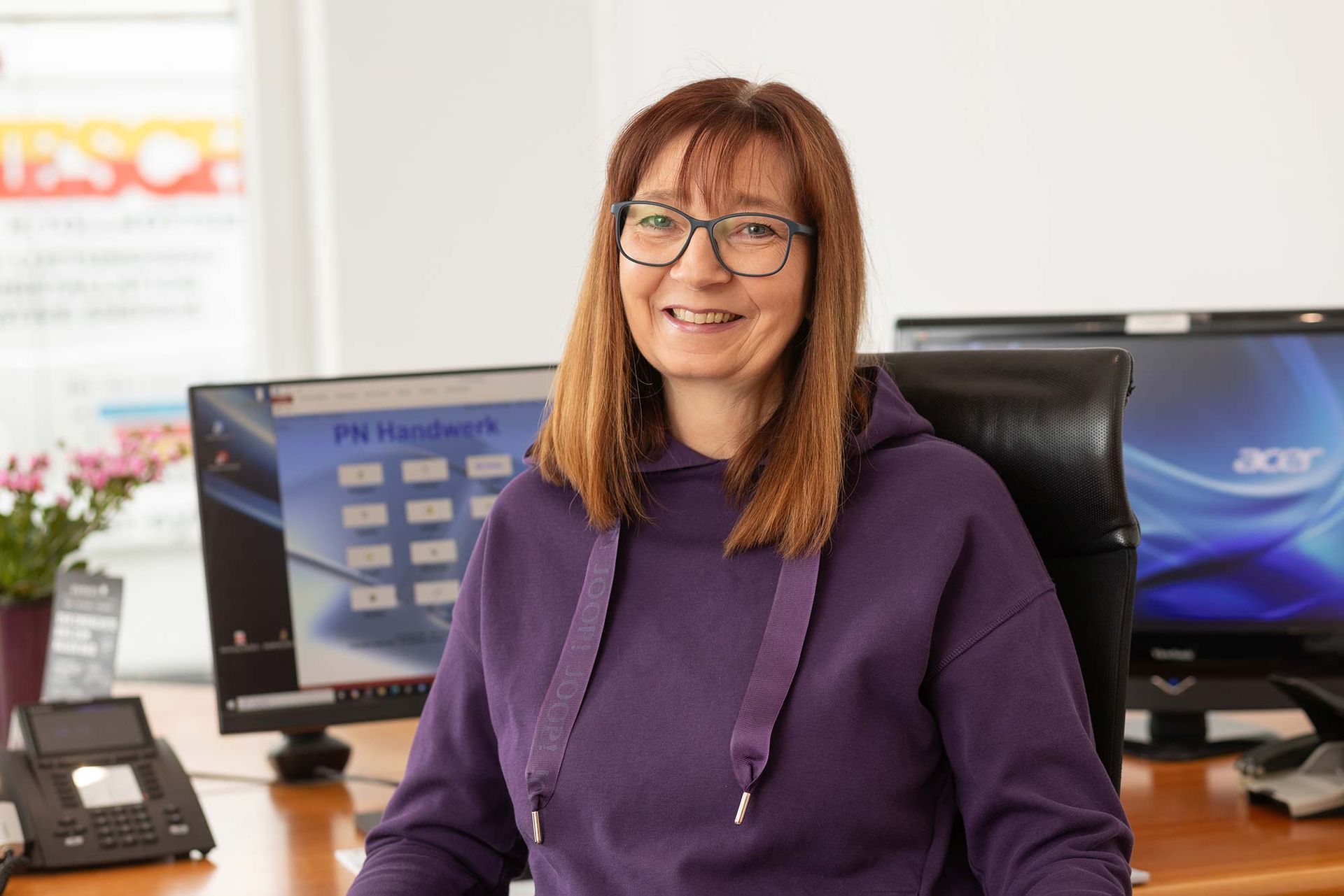 Eine Frau in einem lila Kapuzenpullover sitzt an einem Schreibtisch vor einem Computer.
