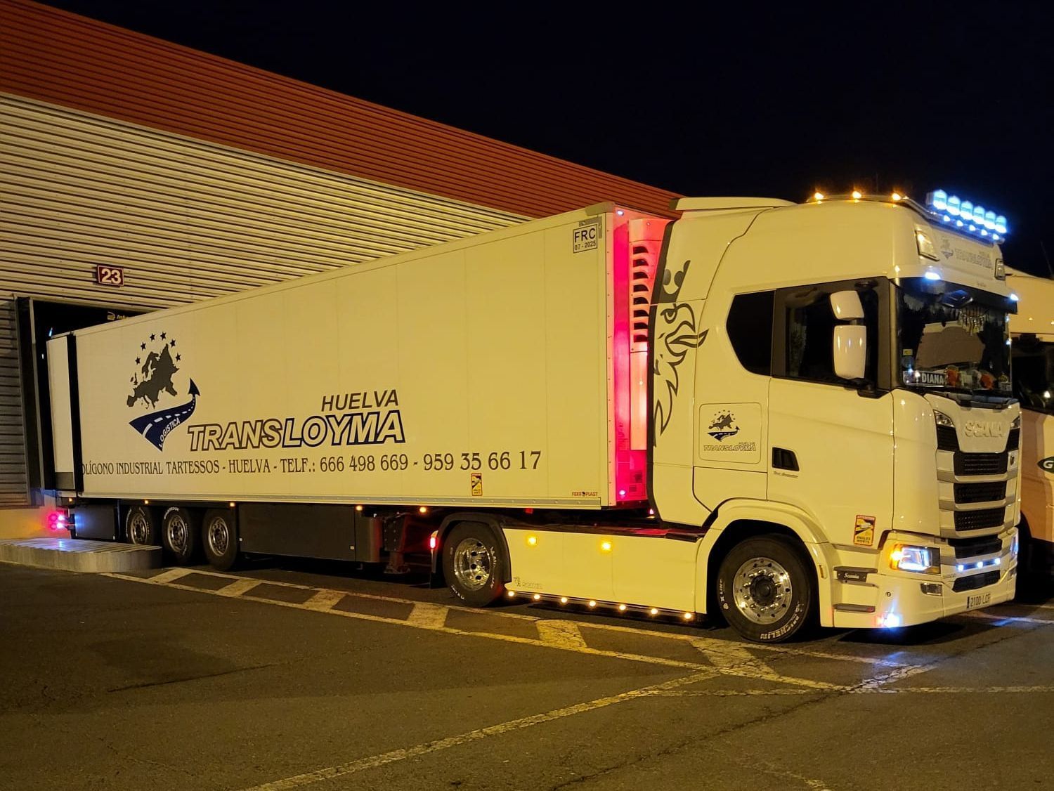 Un camión semirremolque blanco está estacionado frente a un edificio por la noche.