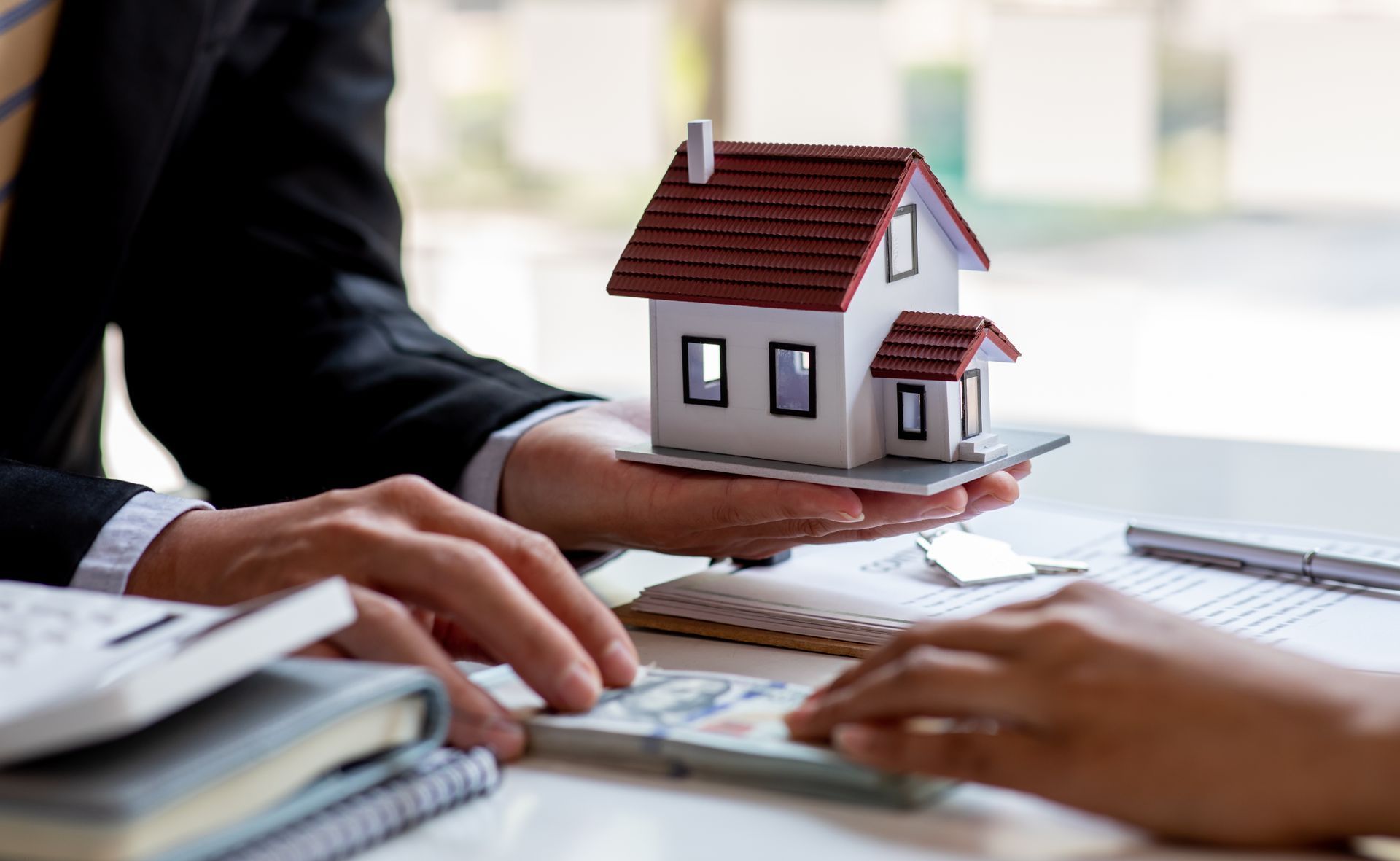 Zwei Personen am Tisch. Eine Person reicht der anderen ein Modellhaus.