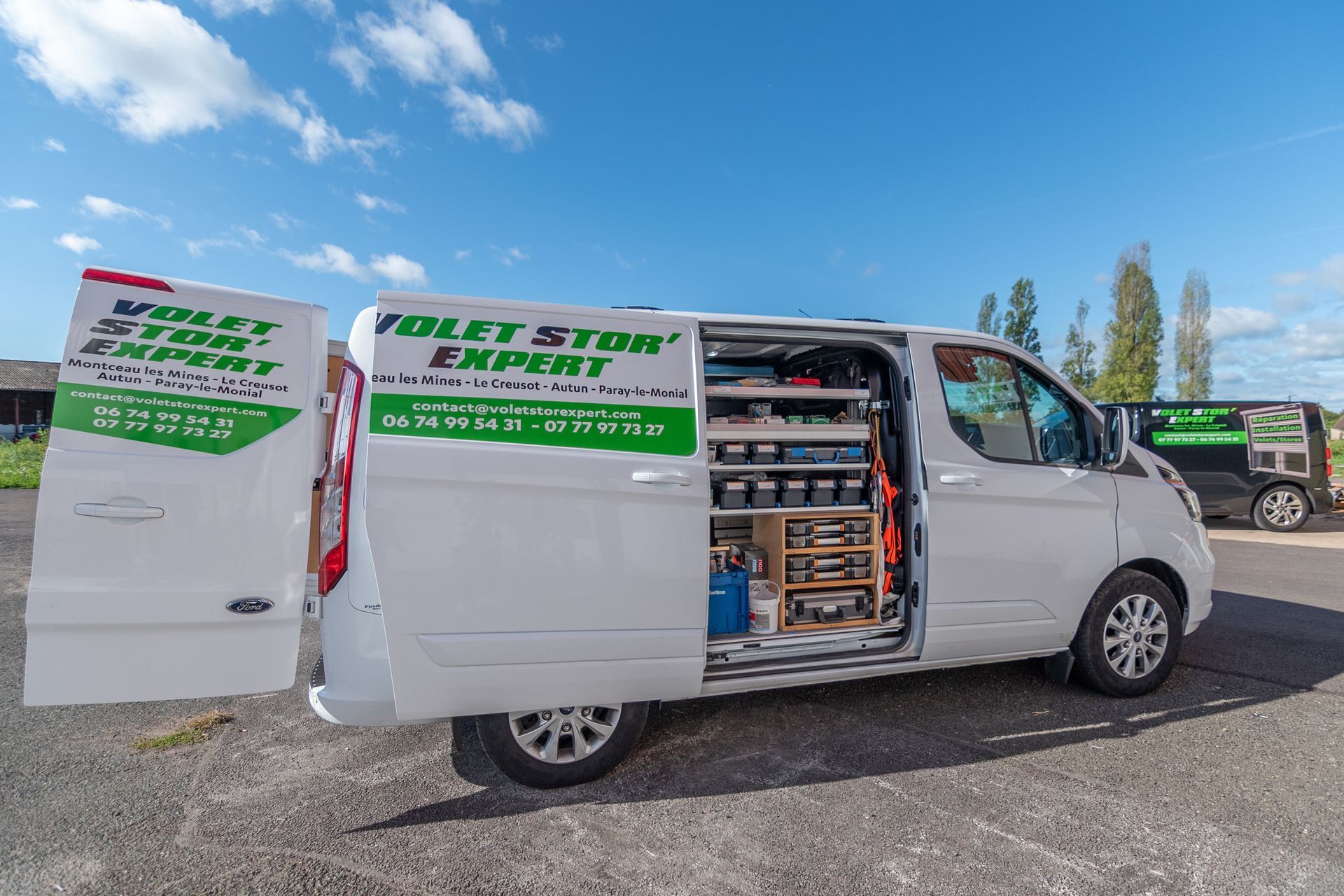 Une camionnette blanche, portes ouvertes, expose des outils, garée sur un parking sous un ciel bleu.