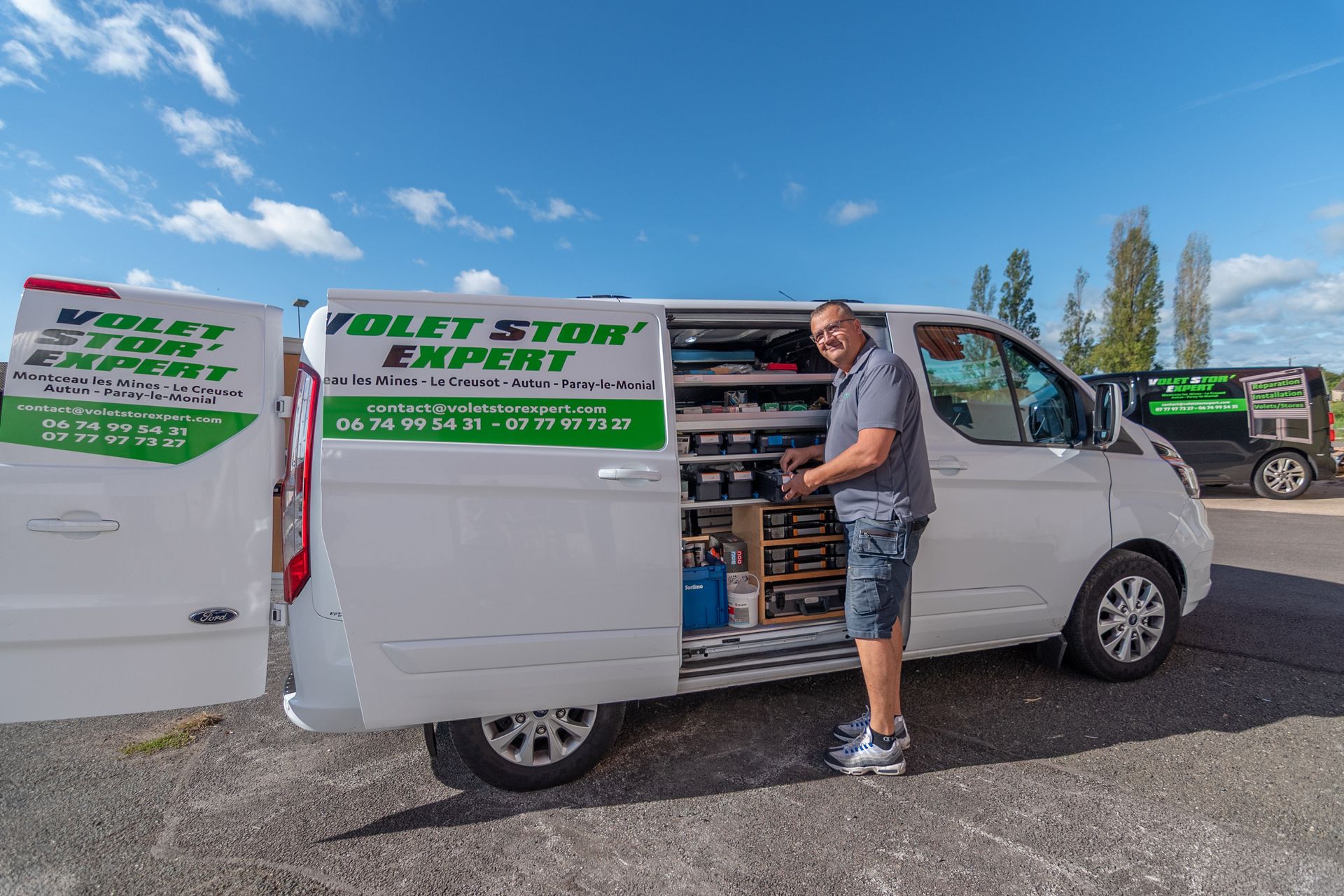 Homme debout près d'une camionnette blanche, portes latérales ouvertes, outils visibles à l'intérieur.