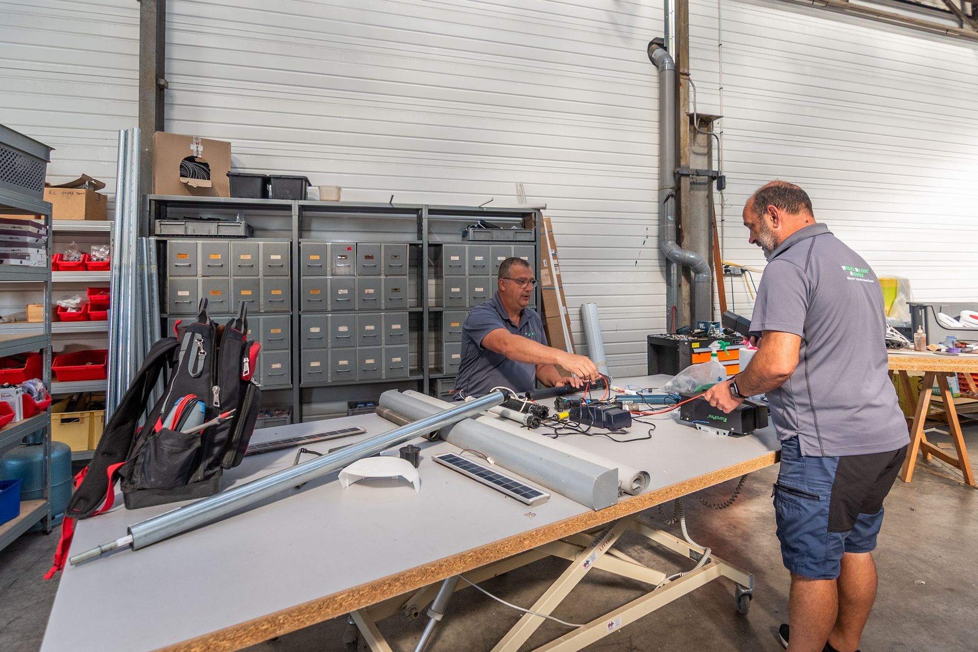 Deux hommes assemblent des pièces sur un établi dans un atelier. Armoires et étagères métalliques avec des pièces en arrière-plan.
