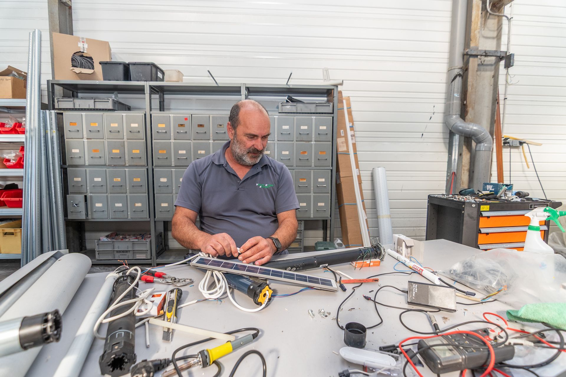 Un homme travaille sur un appareil électronique à un établi. Outils, câbles et pièces sont étalés. Des armoires de rangement en arrière-plan.