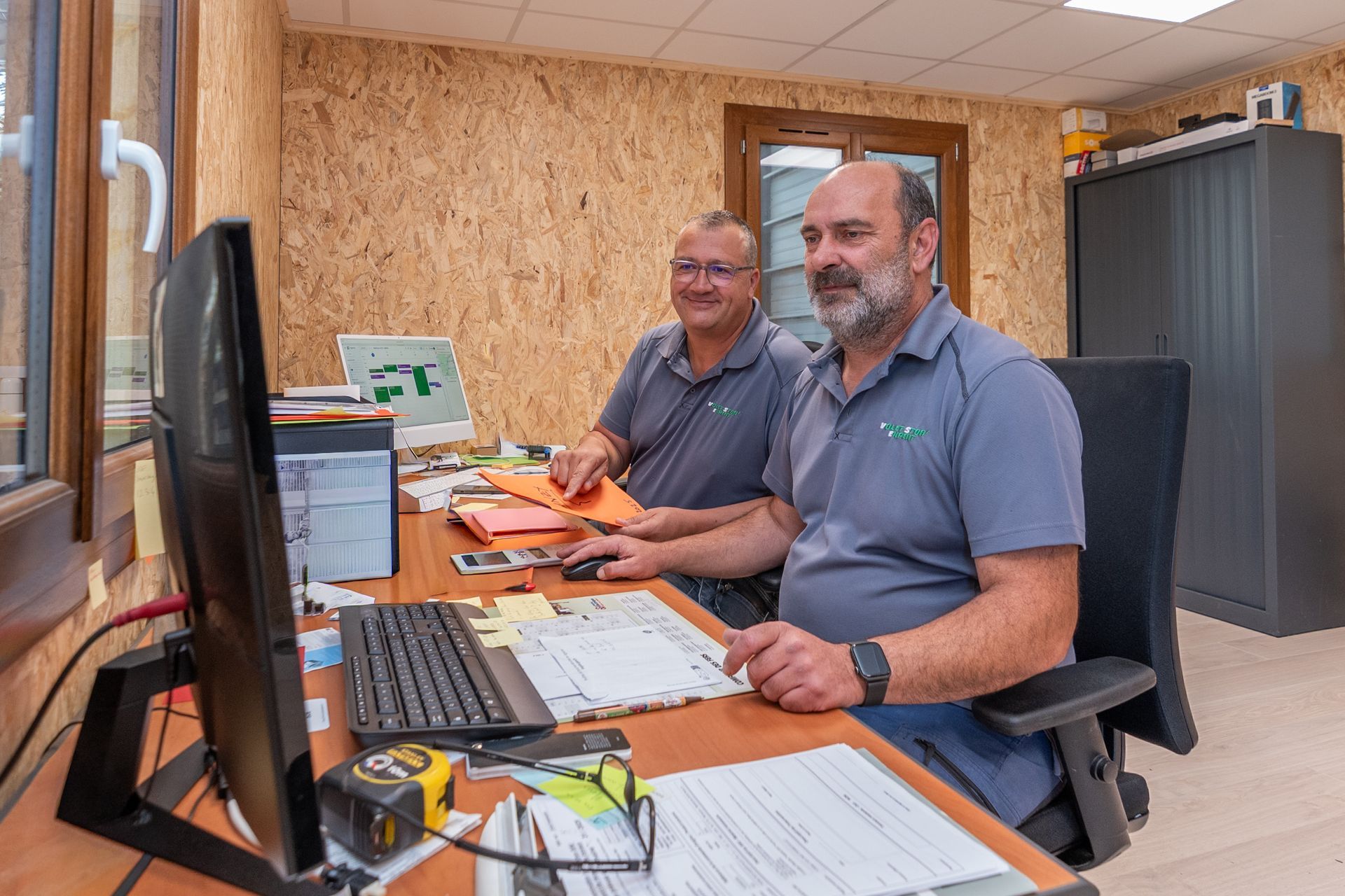 Deux hommes dans un bureau face à un ordinateur, l'un tenant des papiers.