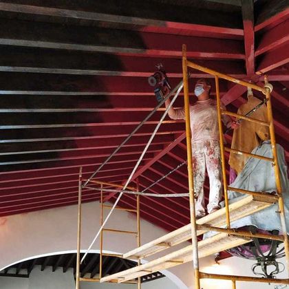 Un trabajador pinta un techo rojo en un andamio amarillo dentro de un edificio.