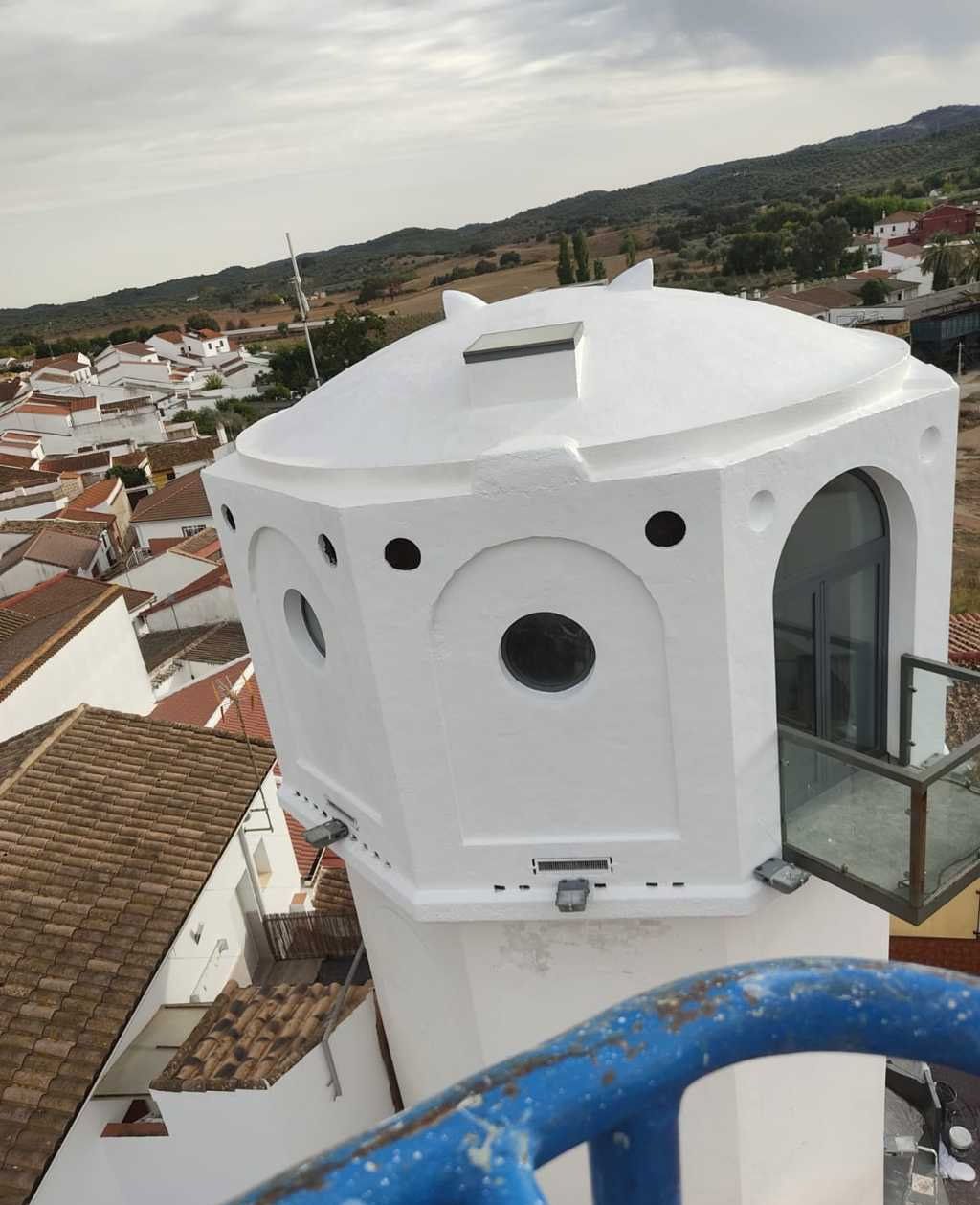 Edificio redondo blanco con ventanas circulares y balcón de cristal con vistas a un pueblo en la ladera de una colina.