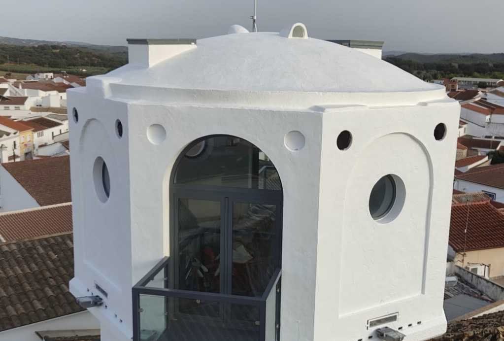 Torre octogonal blanca con techo, ventanas redondas y una puerta de cristal con vistas a un pueblo.