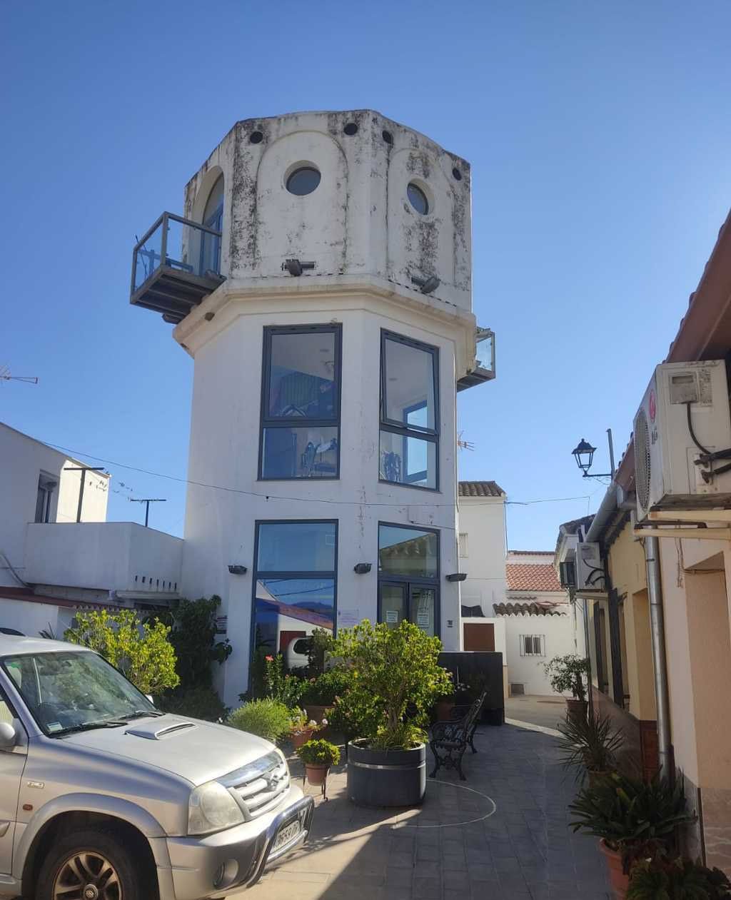 Calle estrecha con un edificio alto y blanco parecido a una torre, un coche aparcado y macetas con plantas bajo un cielo azul.
