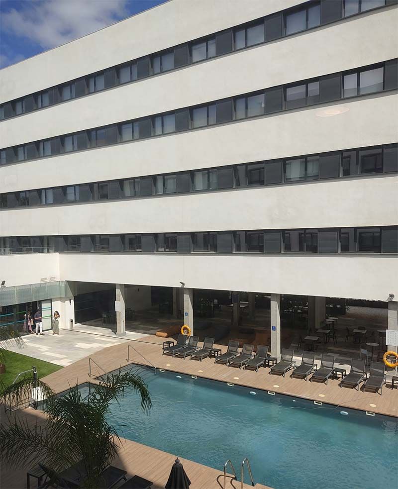 Hotel moderno con una larga fachada blanca que da a una piscina exterior azul y a tumbonas en la planta baja.