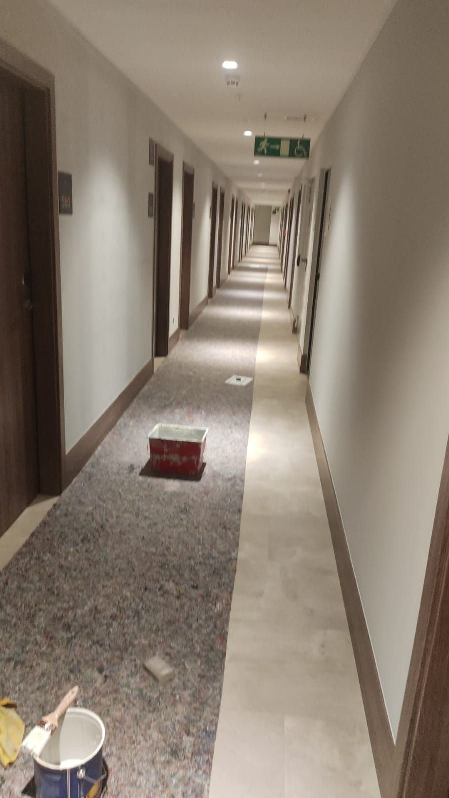 Largo pasillo de hotel con paredes beige, alfombra y un cubo de limpieza en primer plano.