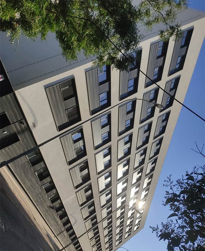 Vista inclinada de un edificio de apartamentos moderno contra un cielo azul, con ramas de árboles en primer plano.