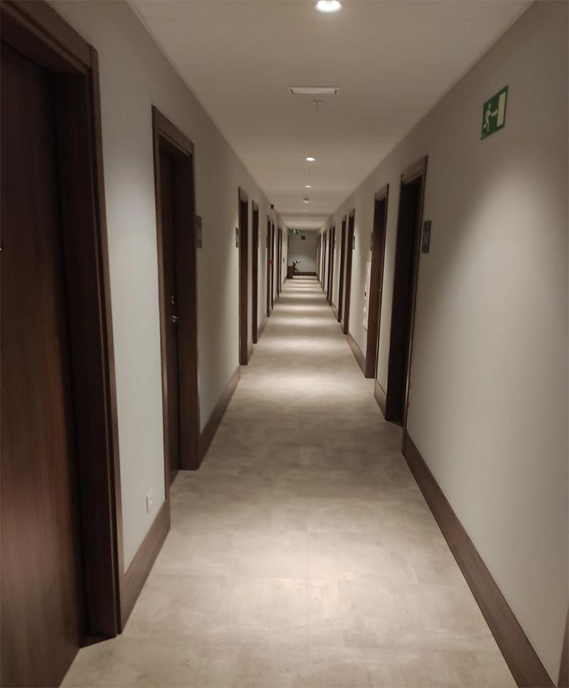 Largo pasillo de hotel con alfombra beige, puertas marrones y luces empotradas en el techo.