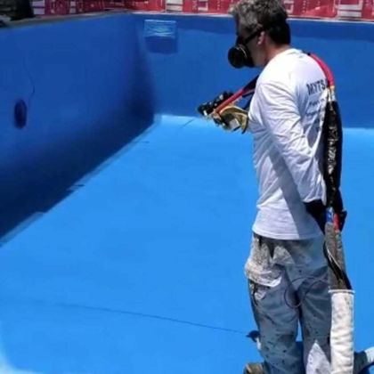 Un trabajador con camisa blanca rocía el interior de una piscina de color azul brillante con una pistola de pintura y un respirador.