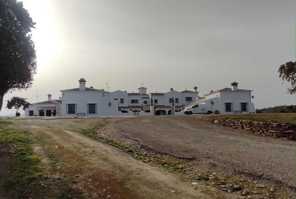 Complejo de edificios blancos junto a un camino de grava en un paisaje rural brumoso.
