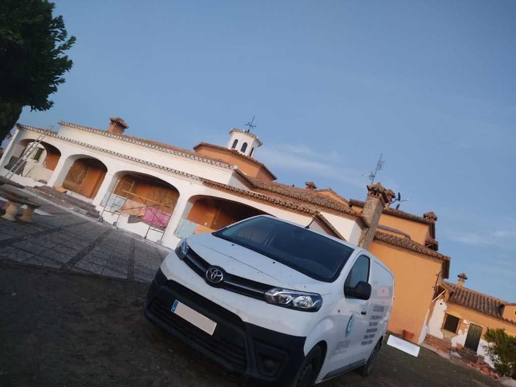 Una furgoneta blanca aparcada frente a una casa soleada de estilo español con porche arqueado y tejado de tejas.