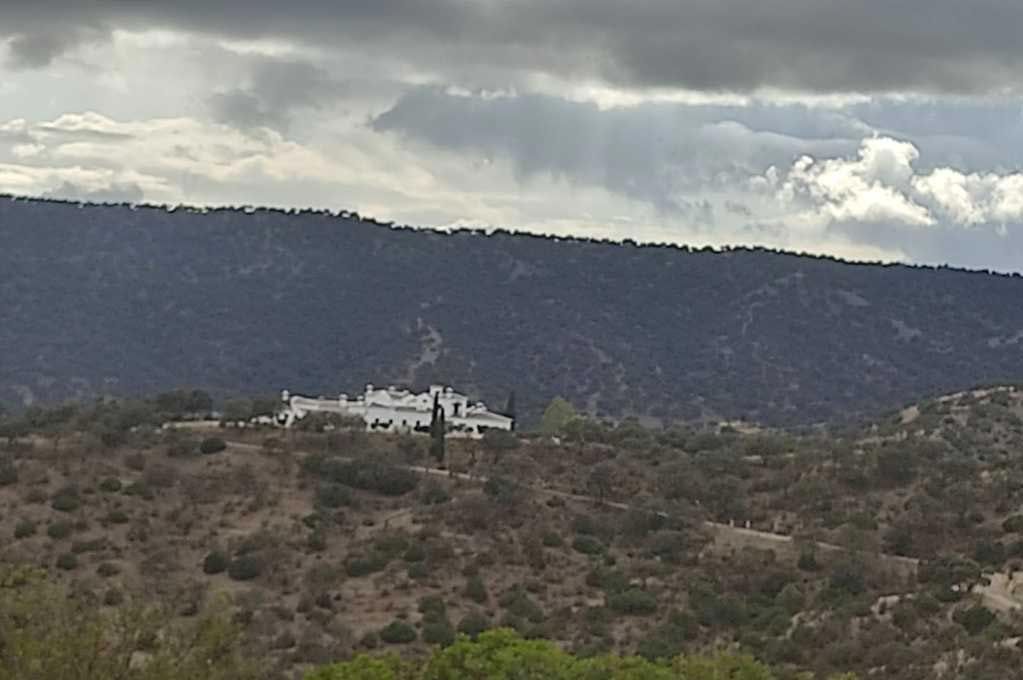 Edificio blanco a lo lejos en una ladera cubierta de matorrales bajo un cielo nublado, con una oscura cresta montañosa detrás.