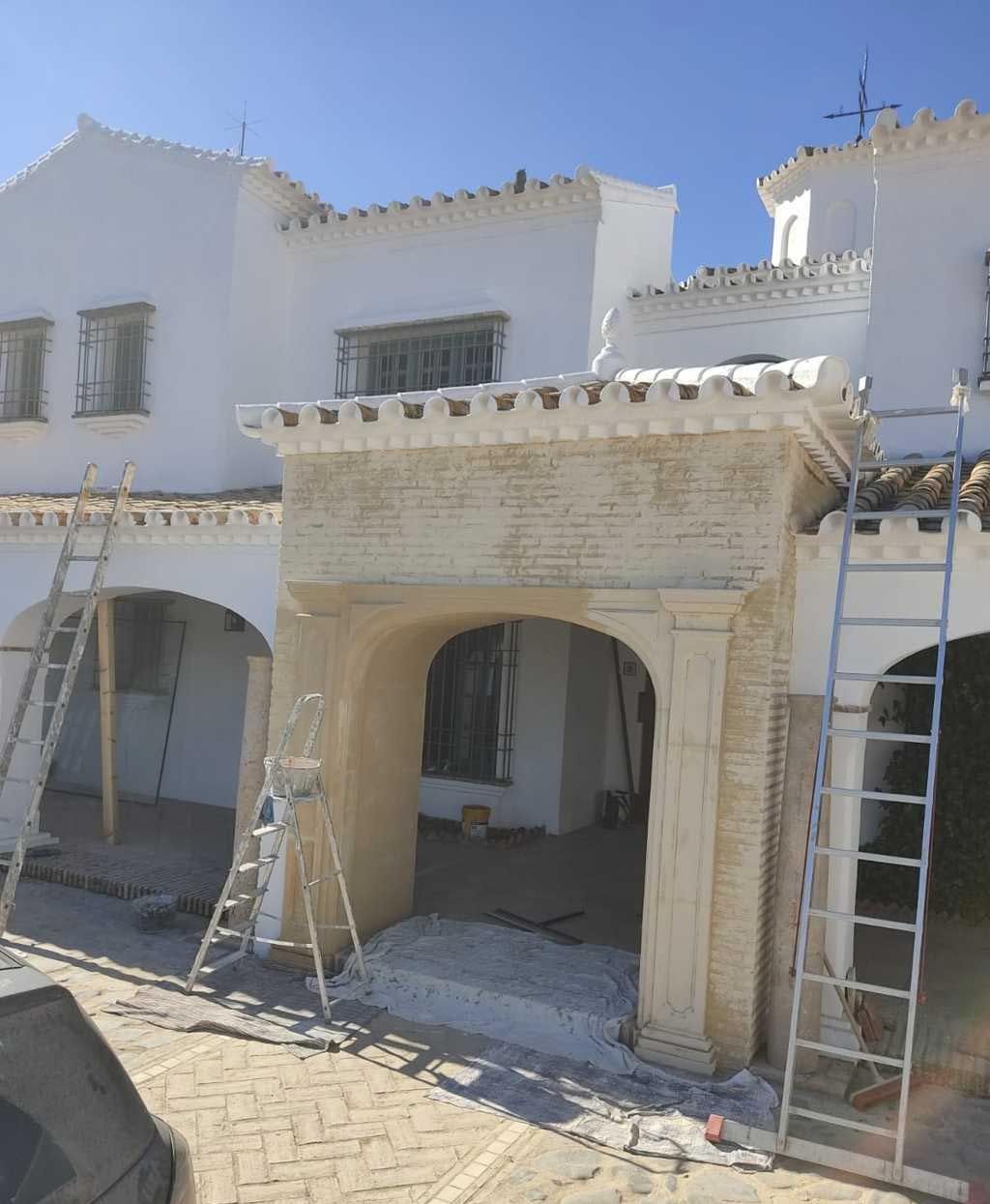 Fachada de una casa de estuco blanco en proceso de renovación, con andamios y escaleras alrededor de las puertas arqueadas.