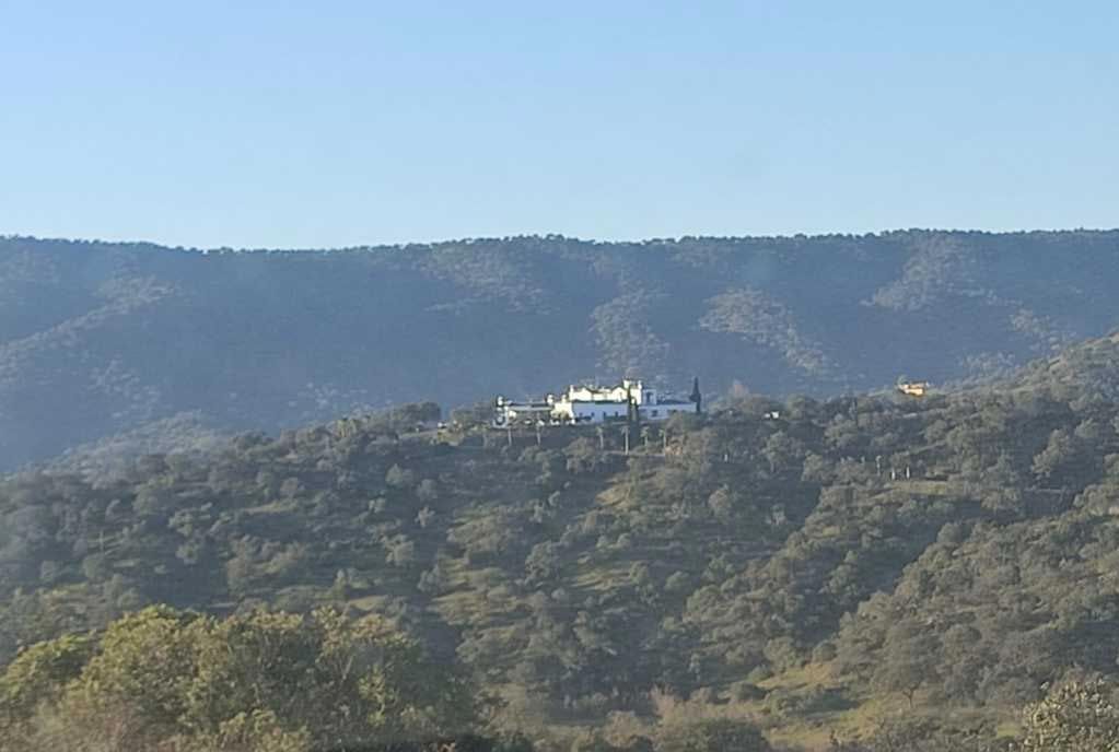 Edificio blanco en una ladera boscosa con montañas al fondo.