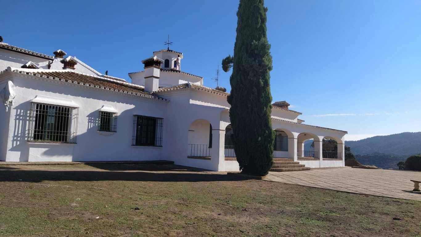 Casa blanca de estilo mediterráneo con contraventanas negras y un alto ciprés en una ladera soleada con montañas en la lejanía.