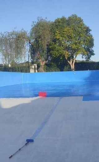 Un terreno cubierto con una lona azul, con un cubo rojo y una herramienta de mango largo delante de una valla azul y árboles.