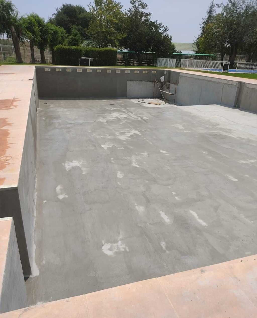 Piscina de hormigón vacía en construcción, rodeada de muros bajos y con árboles al fondo.