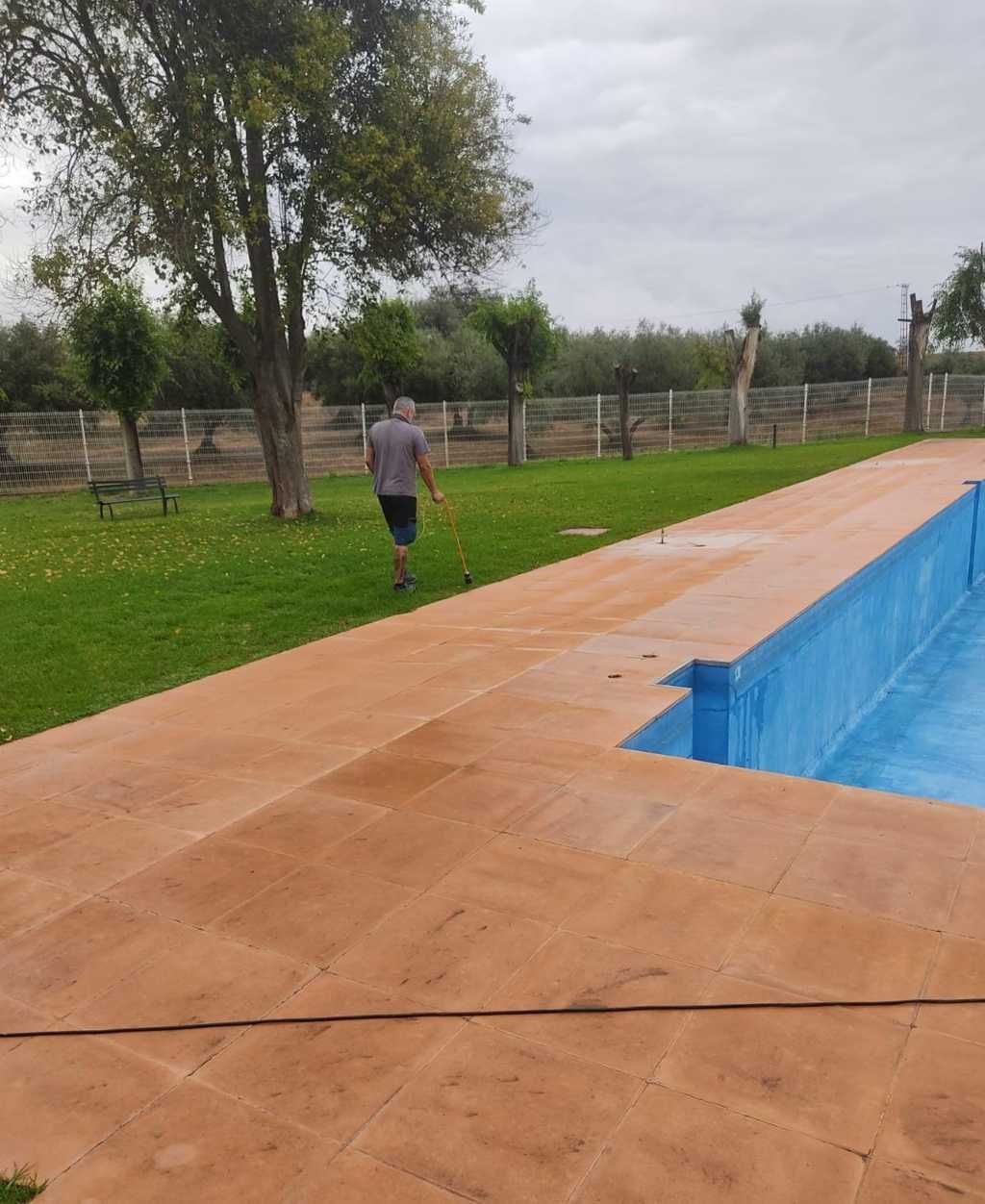 Persona caminando junto a una piscina al aire libre con una terraza de madera y una zona de césped al fondo.