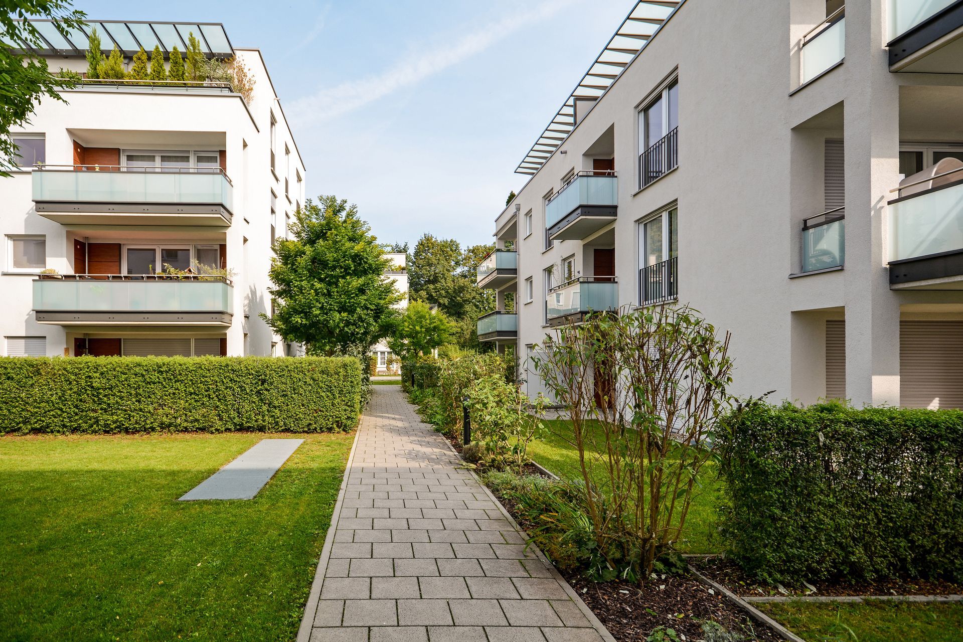 Une allée pavée relie deux immeubles d'appartements modernes aux couleurs claires, entourés de pelouses verdoyantes et de haies.