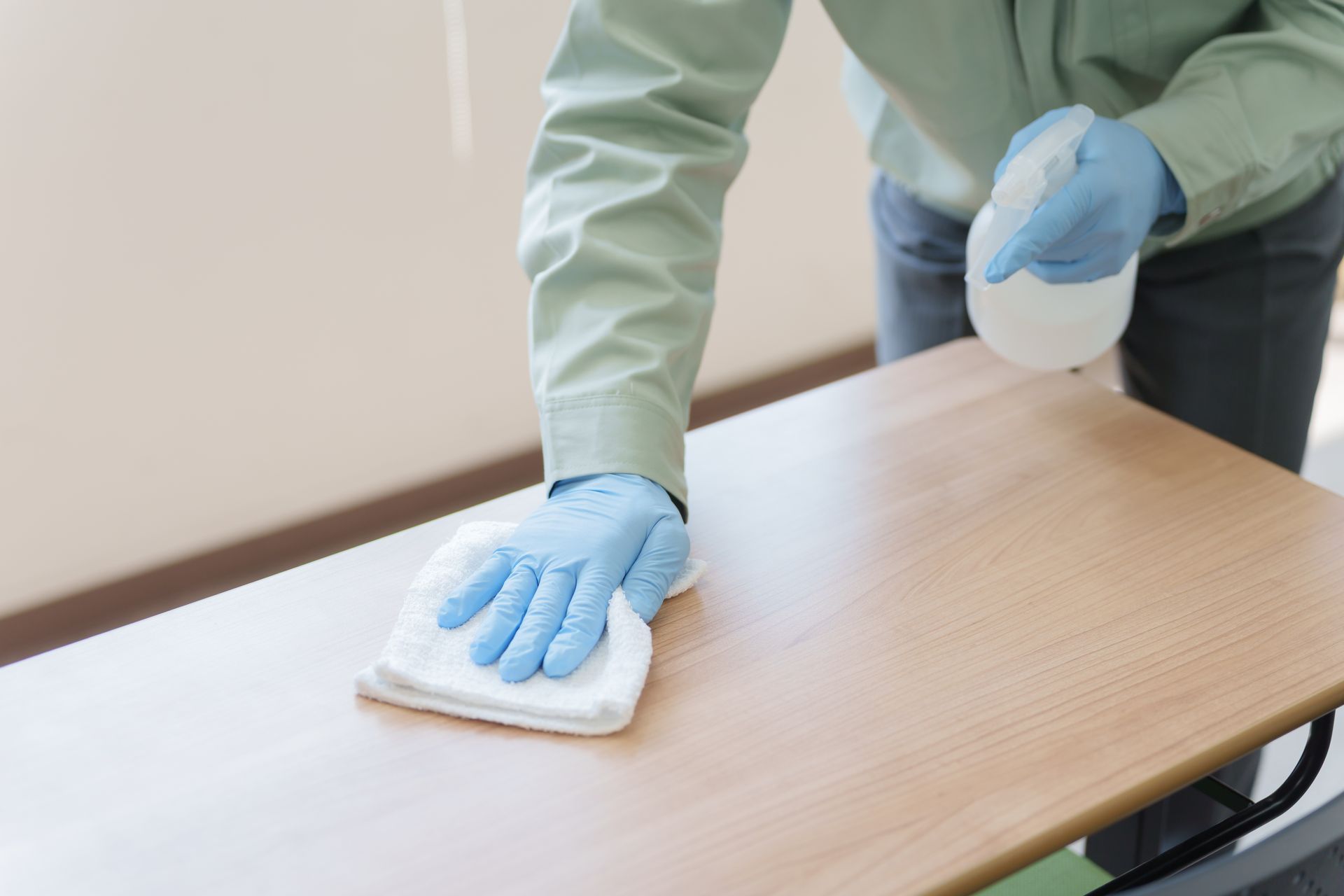 Une personne portant des gants bleus nettoie une table en bois avec un chiffon blanc tout en tenant un flacon pulvérisateur.