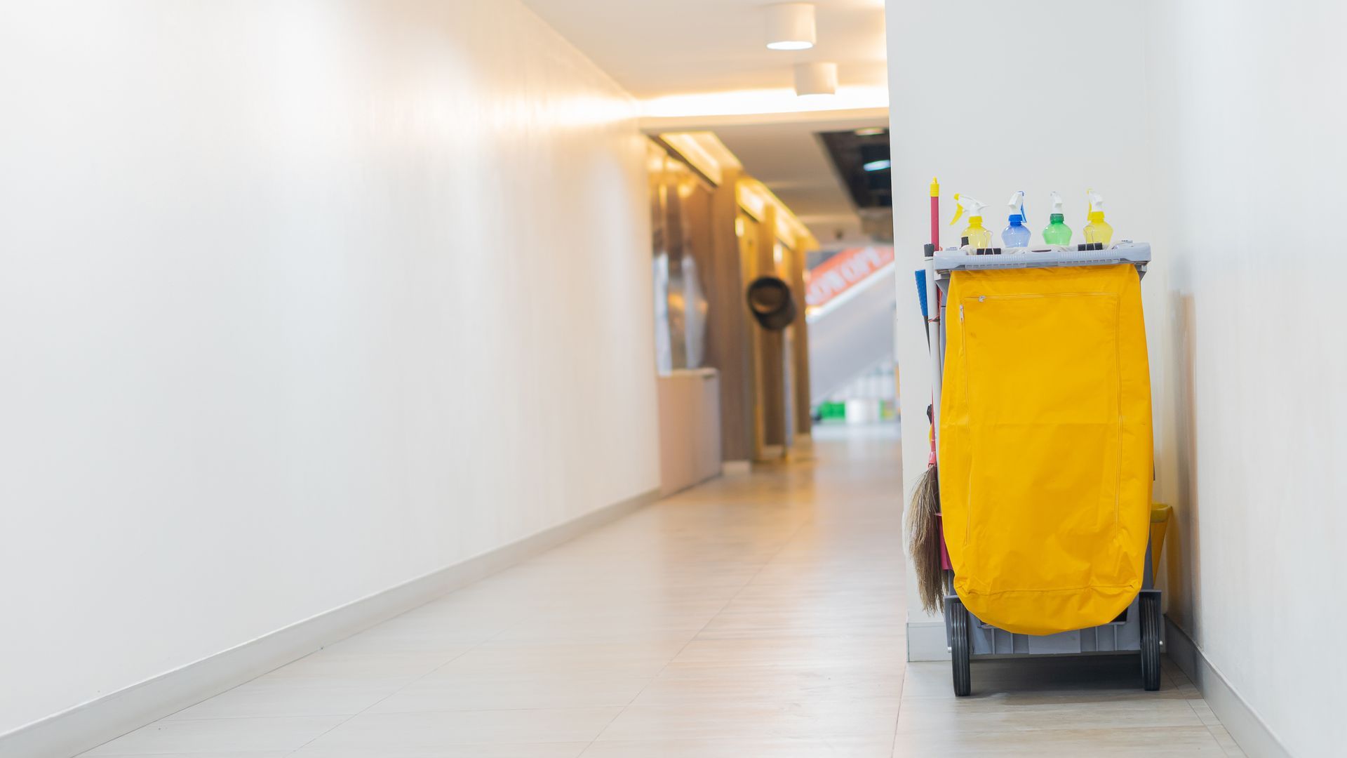 Un chariot de nettoyage jaune se trouve dans un couloir blanc, calme et bien éclairé.
