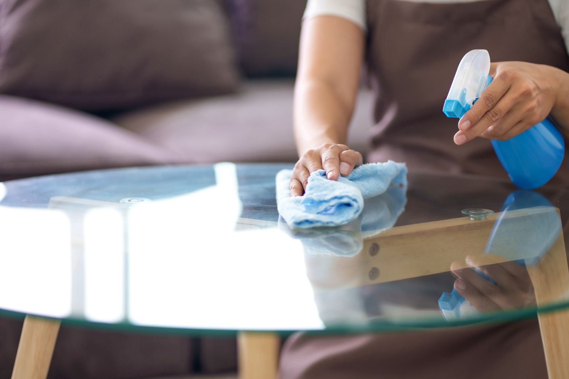 Une personne portant un tablier utilise un flacon pulvérisateur bleu et un chiffon pour nettoyer une table basse ronde en verre.
