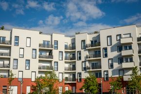 Un immeuble d'appartements moderne à plusieurs étages, avec des murs extérieurs blancs et rouges, des balcons et des fenêtres rectangulaires sous un ciel bleu.