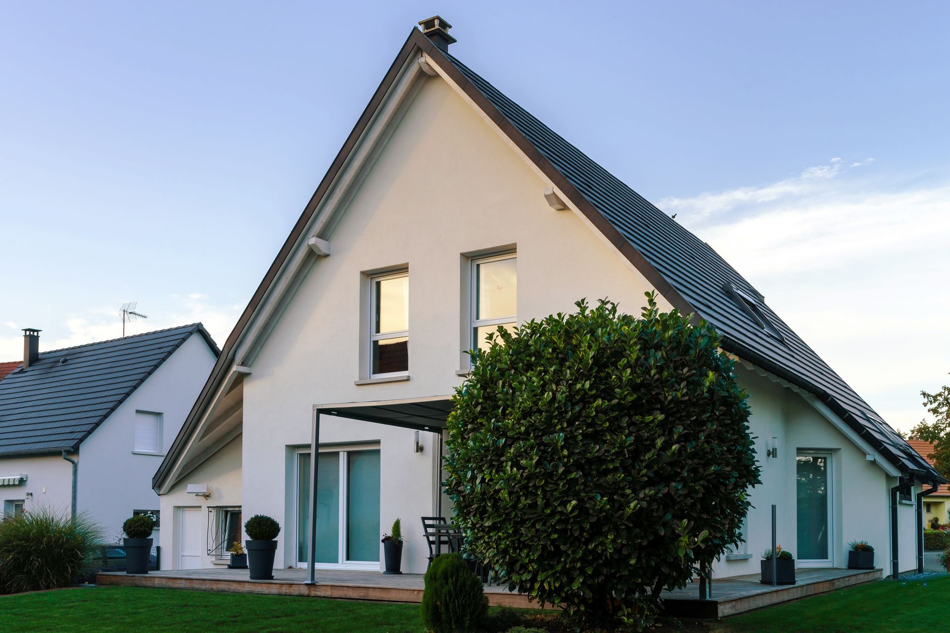 Une maison blanche à deux étages avec un toit de tuiles grises à forte pente, une pelouse et un grand arbuste rond dans la cour avant.