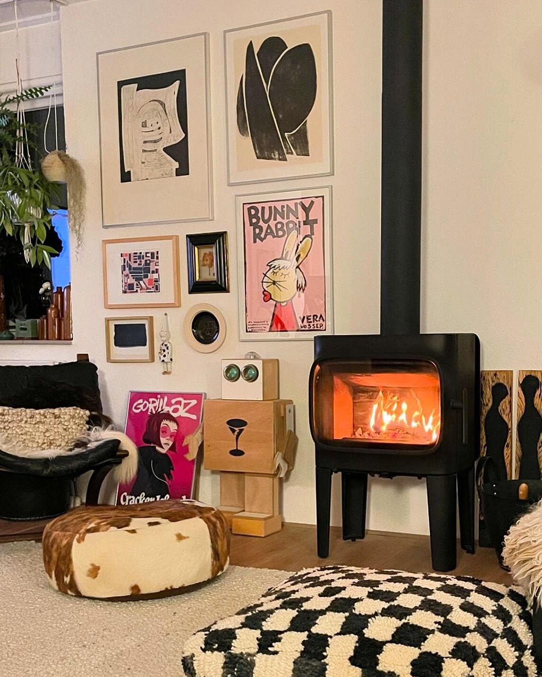 Salon cosy avec poêle à bois, reproductions d'art au mur, poufs en cuir et coussin de sol à carreaux noirs et blancs.