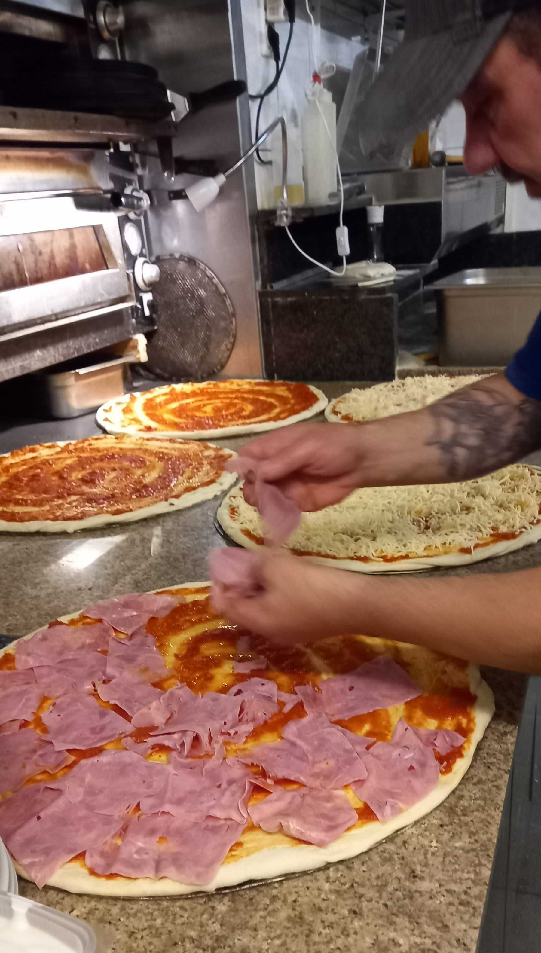 Ein Mann legt in einer Küche Schinken auf eine Pizza, Leonardo Intelisano Pizzeria Dolce Vita