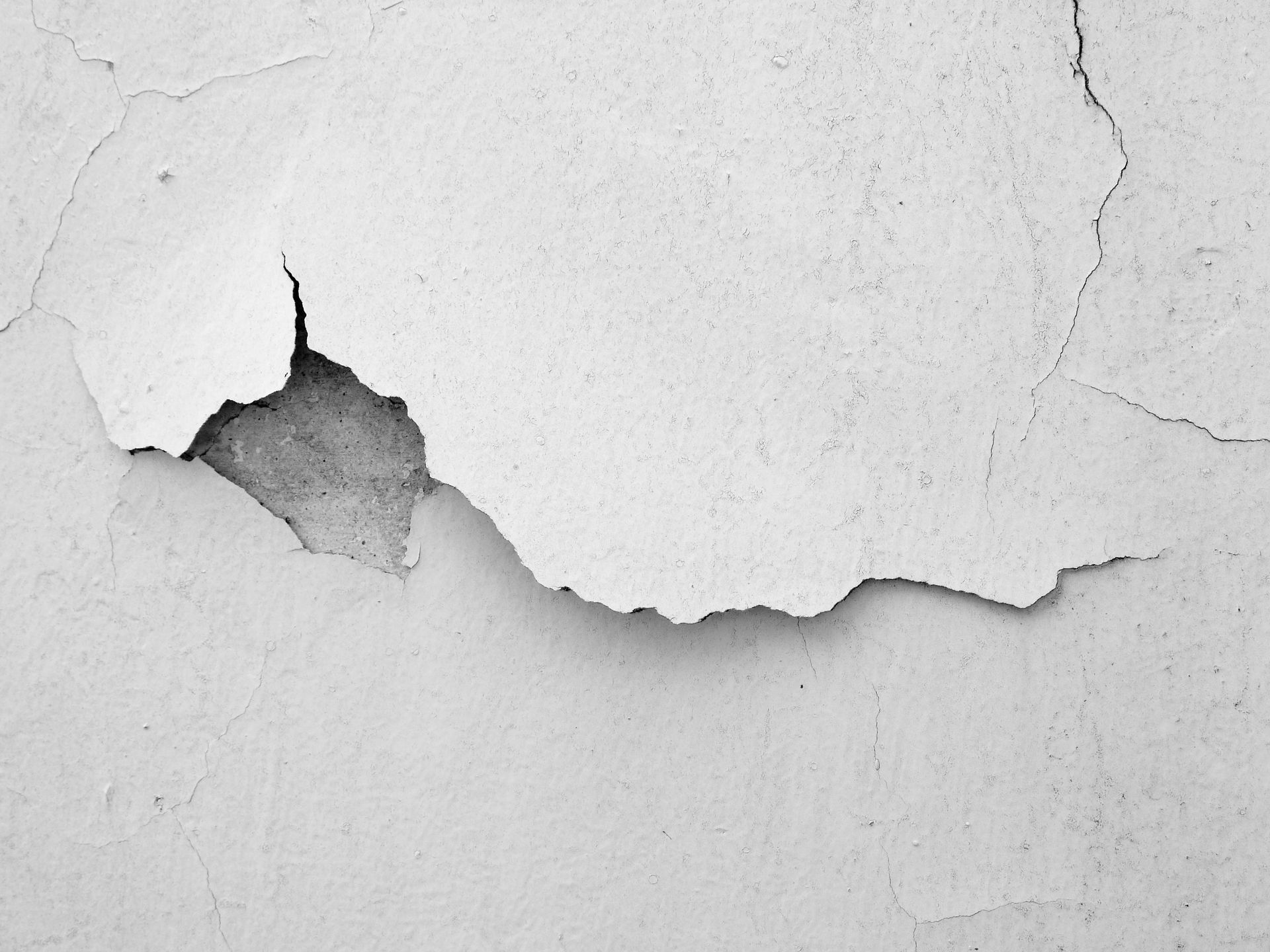 Peinture blanche craquelée et écaillée sur un mur texturé, révélant le béton gris rugueux en dessous.