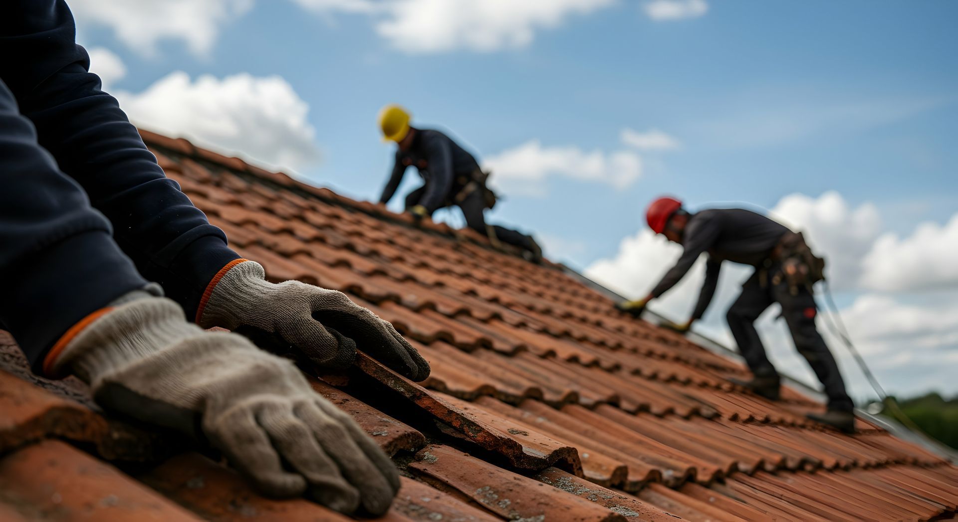 Des couvreurs en équipement de sécurité travaillent sur un toit en tuiles en terre cuite sous un ciel nuageux.