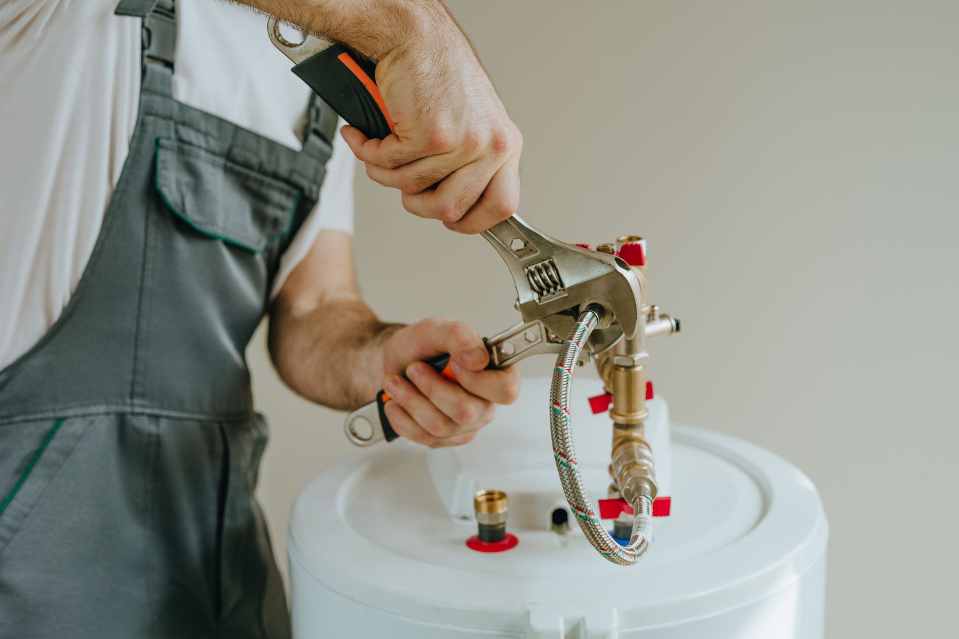 Plombier en salopette grise utilisant une clé pour travailler sur un chauffe-eau.
