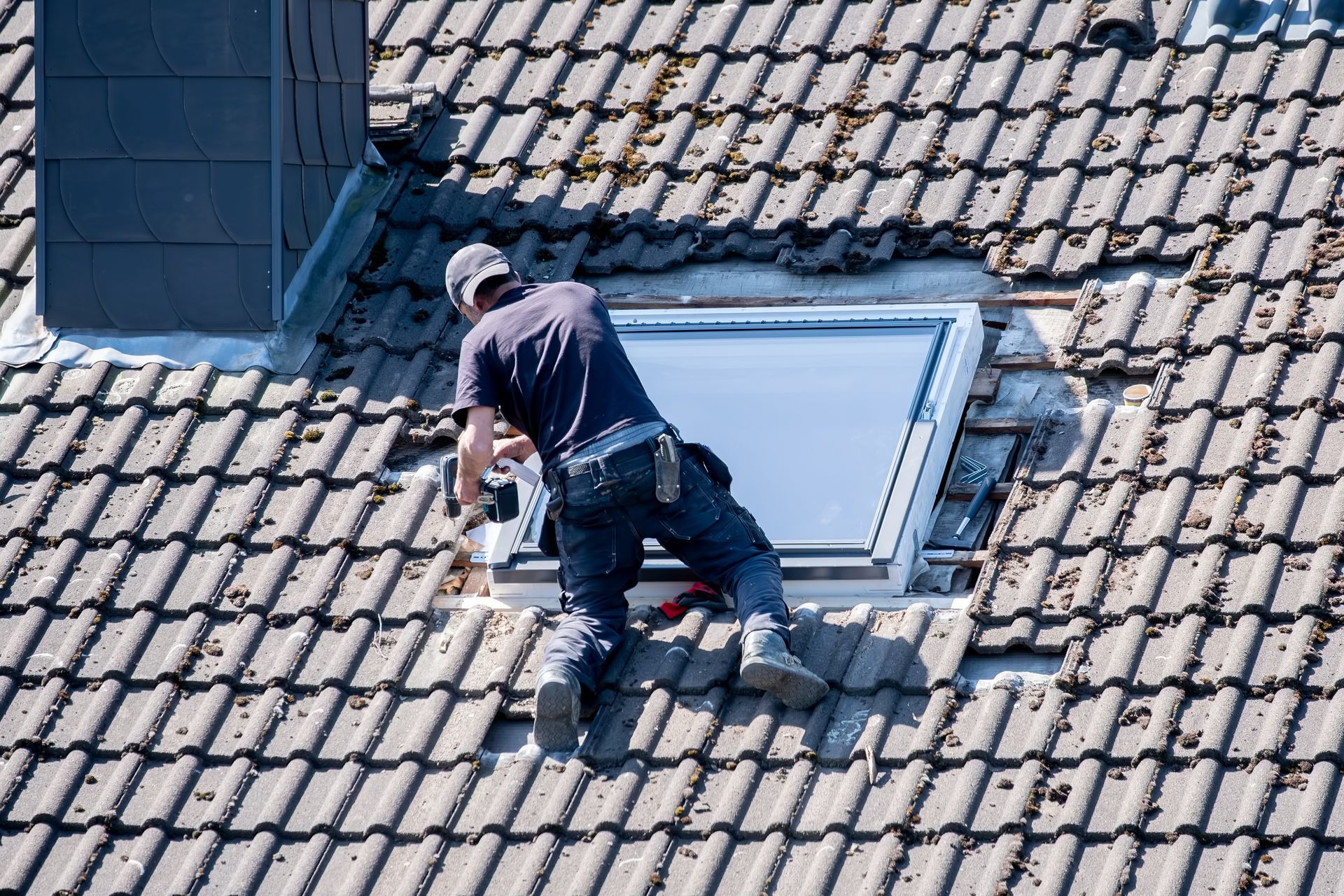 Couvreur sur un toit en tuiles, installant une lucarne. Il porte une casquette, un jean et utilise un outil.