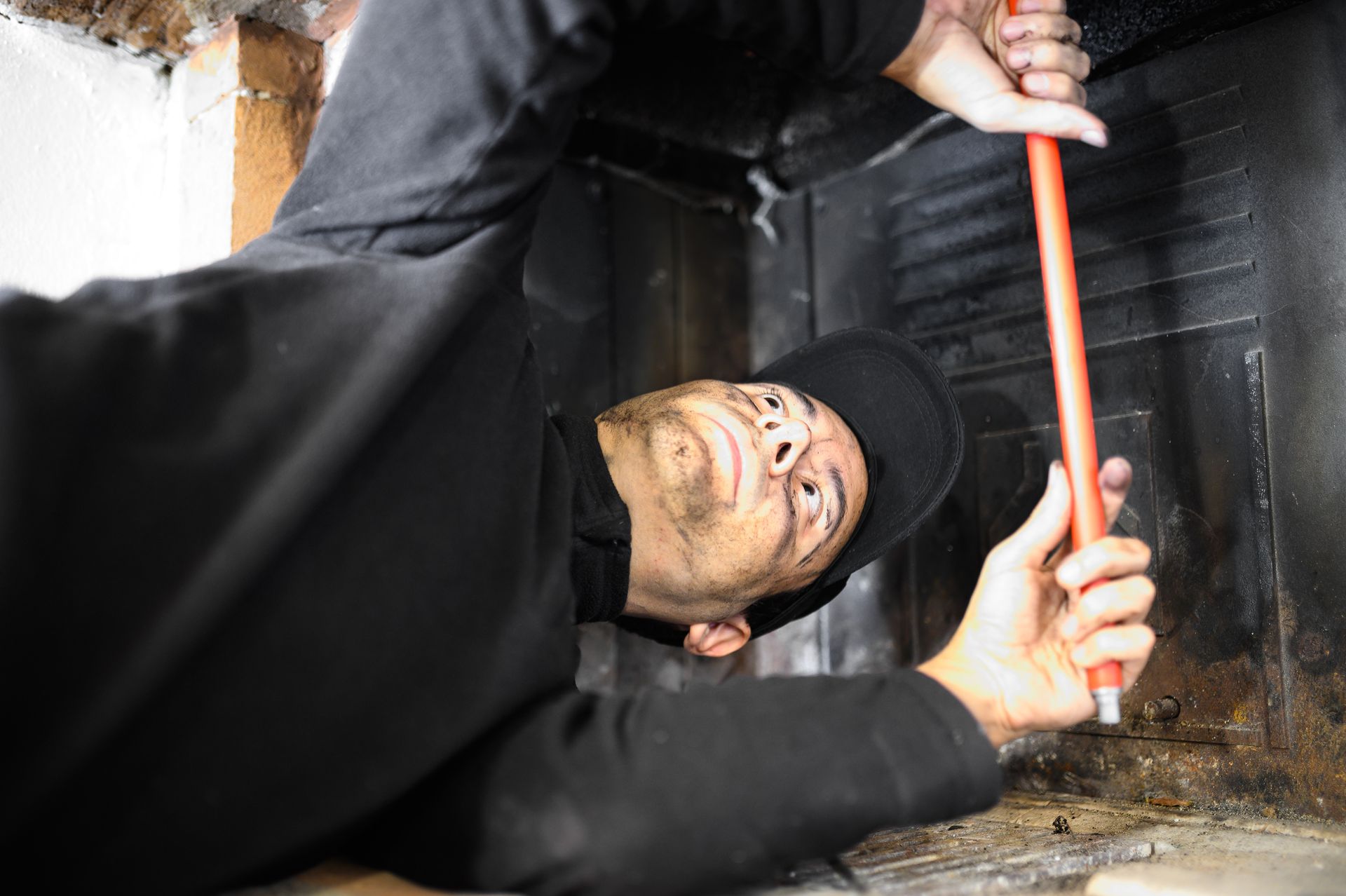 Homme nettoyant une cheminée, suie sur le visage, vêtements noirs, utilisant une longue perche rouge.