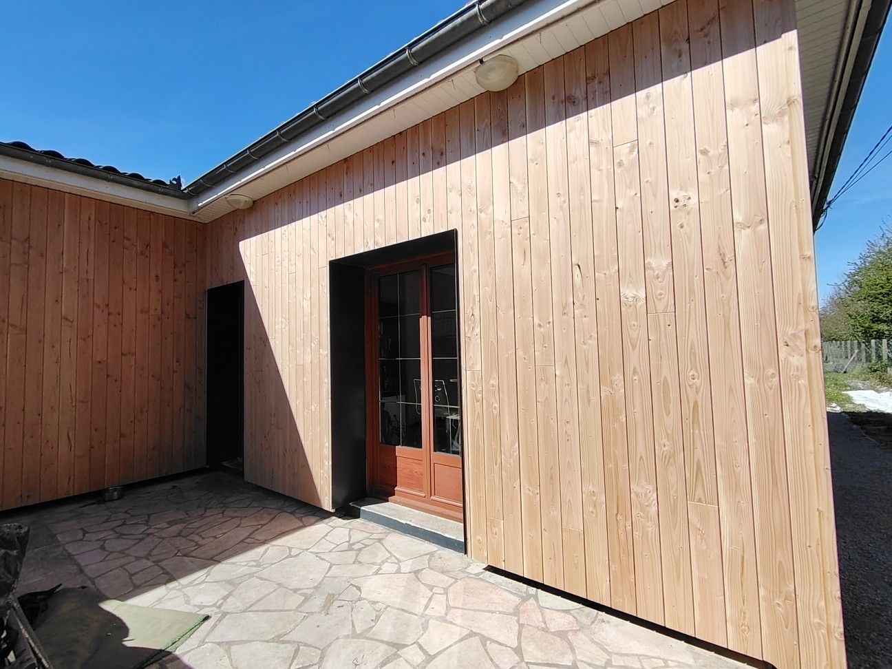Maison à façade en bois avec porte vitrée et terrasse en pierre sous un ciel bleu