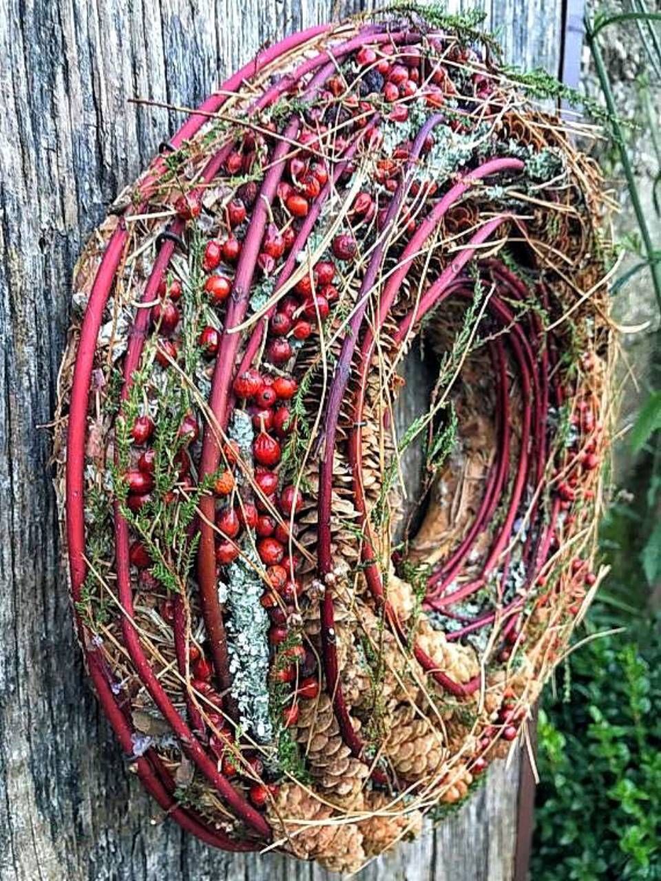 Ein Kranz aus roten Zweigen, Beeren und Tannenzapfen hängt an einer Holzfläche.