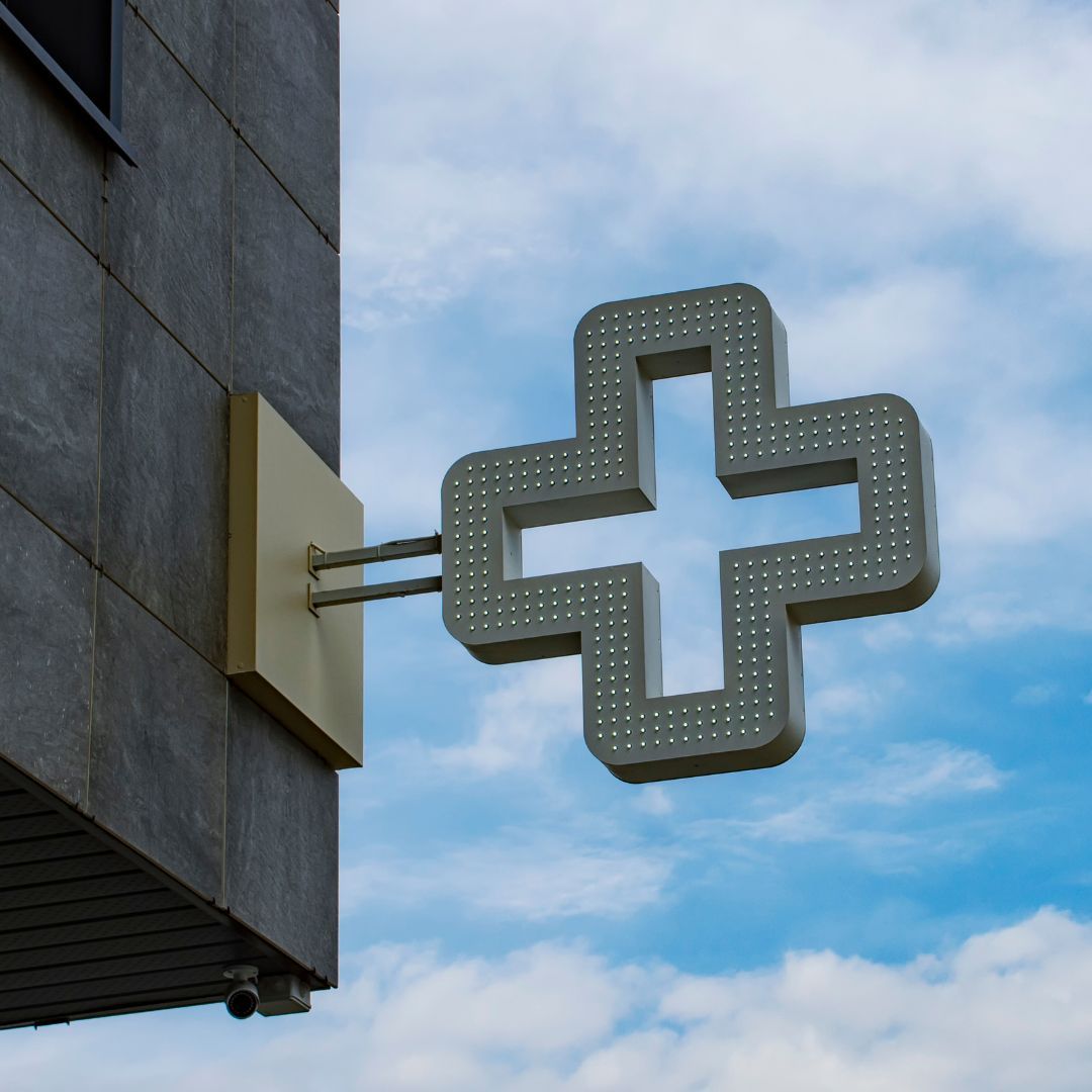Un cartello metallico a forma di croce per farmacia montato sul lato di un edificio in pietra grigia, contro un cielo azzurro con nuvole.