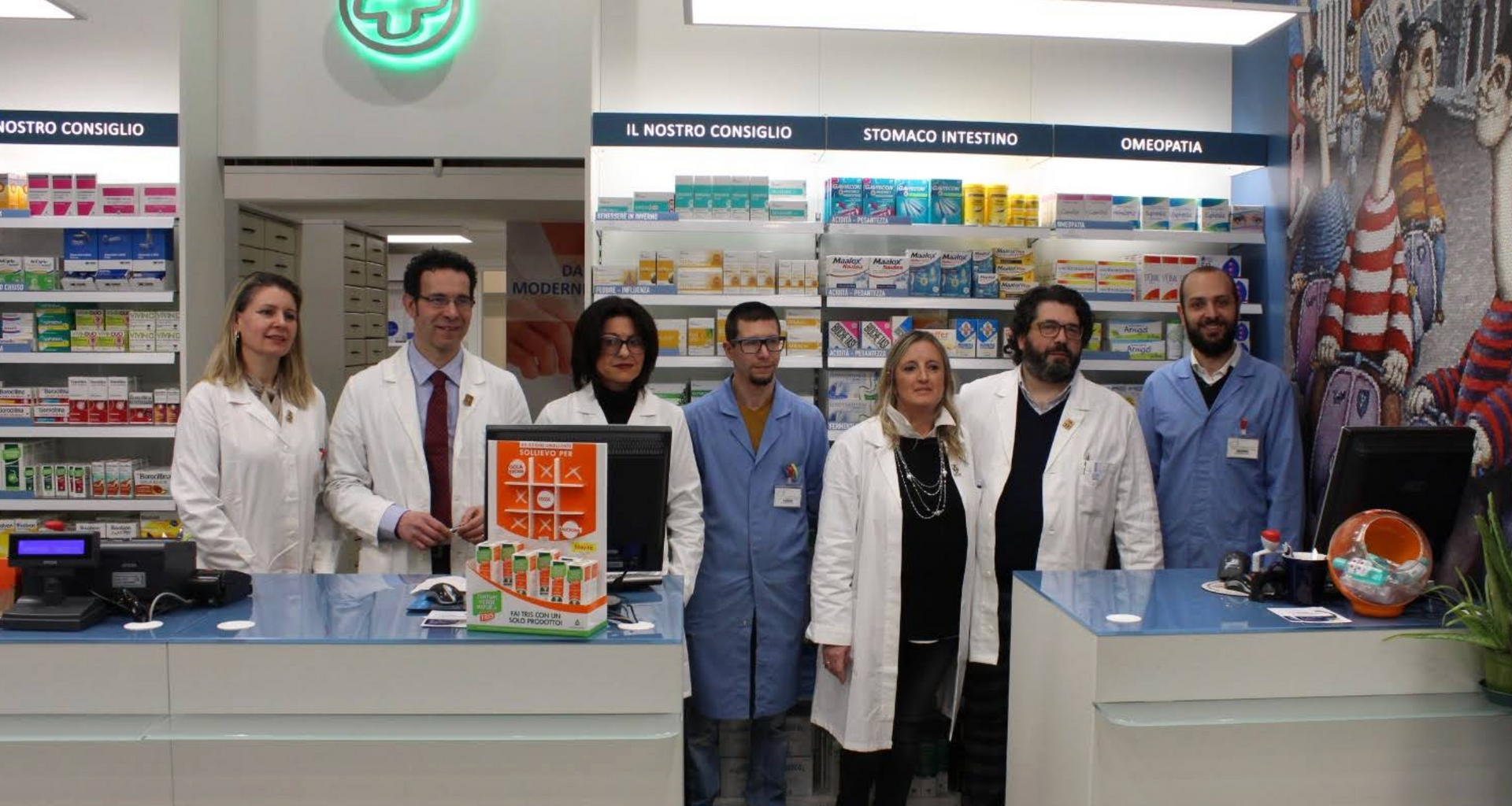 Sette farmacisti in camice bianco e uniforme blu in fila dietro un bancone.