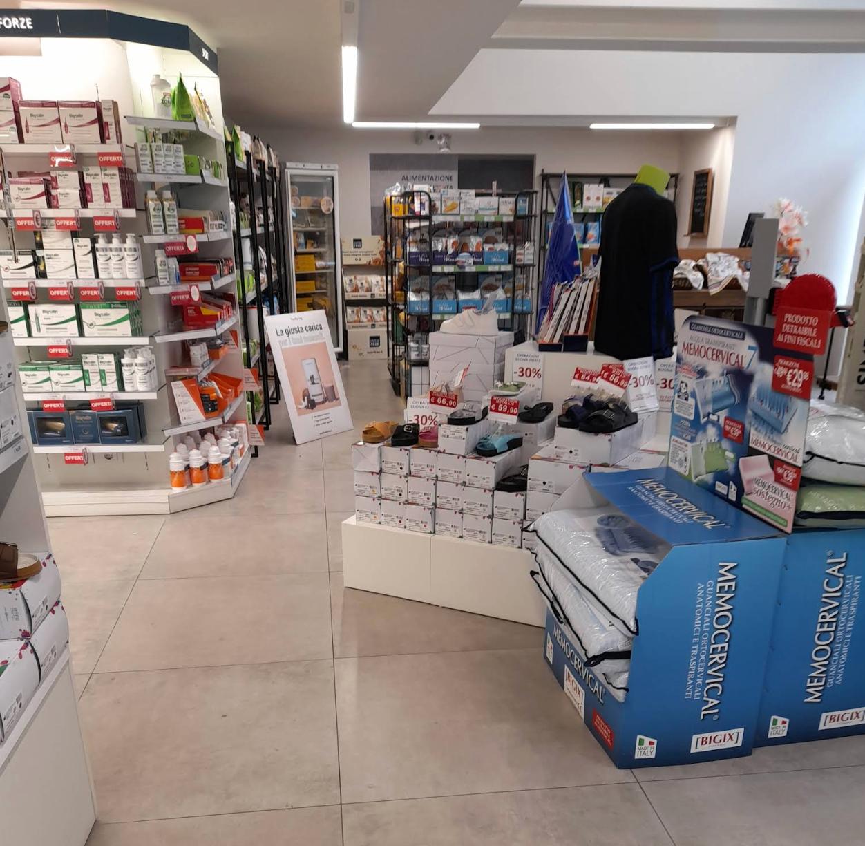 Interno di una corsia di un negozio con scaffali di prodotti sanitari, un'esposizione di scatole e una persona in piedi sullo sfondo.