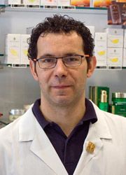 Una persona con camice bianco e occhiali da laboratorio, in piedi davanti a scaffali pieni di vari prodotti medicinali.