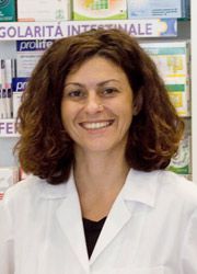 Una persona sorridente con indosso un camice bianco da laboratorio, davanti a scaffali pieni di prodotti farmaceutici.