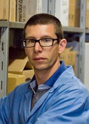 Una persona con i capelli corti e gli occhiali, che indossa una camicia blu con colletto, è in piedi davanti a degli scaffali pieni di scatole.