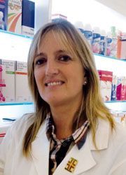 Un farmacista in camice bianco sorride, in piedi in una farmacia con scaffali pieni di prodotti sullo sfondo.