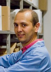 Una persona che indossa un camice blu e una camicia rosa con colletto sorride mentre è in piedi davanti agli scaffali in un magazzino.